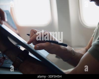 Person using a tablet and stylus on an airplane seat, representing mobile creativity and work during travel. Stock Photo