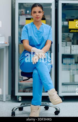 A medical professional is seated in a clinical environment, wearing blue doctor and gloves, ready to assist with patient care or medical tasks in a healthcare facility || Model released Stock Photo