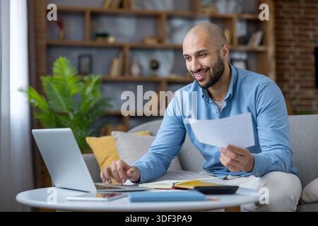 Happy bald man in blue shirt sits on sofa in living room, smiling as he reviews documents and types on a laptop, managing finances and remote work wit Stock Photo