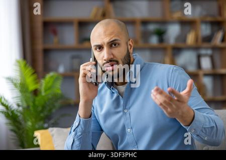 Young bald man making phone call, experiencing communication issues and expressing frustration or misunderstanding while teleworking from a home offic Stock Photo