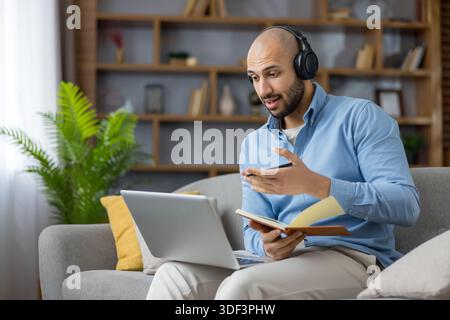 Young man wearing headphones and holding a notebook having a video conference on a laptop from home, actively participating in remote work or online e Stock Photo
