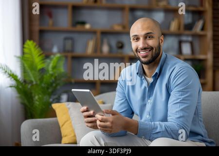 Happy young man relaxing on a modern sofa in his living room, smiling at camera while browsing a tablet casual, connected lifestyle, remote work and d Stock Photo