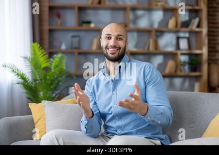 Happy young man sitting comfortably on a gray sofa in a modern living room, smiling at the camera and actively gesturing with his hands while communic Stock Photo