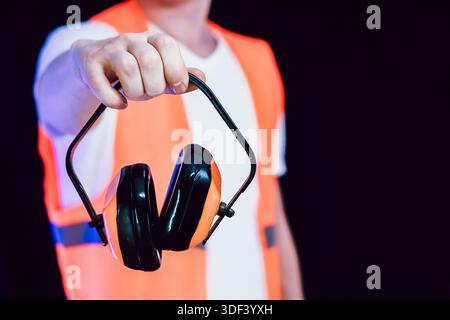 Close-up of hand in orange safety vest clutching black and orange noise protection earmuffs against dark backdrop Stock Photo