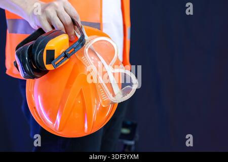 Close-up of safety gear: orange helmet with attached yellow earmuffs and clear protective goggles held by worker on black background. Stock Photo