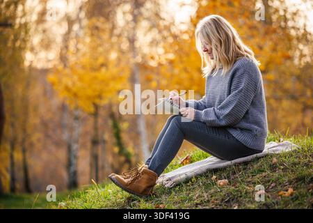 Relaxed woman is writing her diary and resting in autumn park. Adult woman practicing mindfulness and slow living through journaling Stock Photo