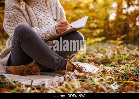 Woman sitting on blanket in autumn forest writing diary. Journaling for good mental wellbeing and mindfulness intentions Stock Photo