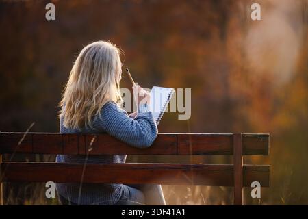 Thoughtful woman sitting on park bench and writing diary or inspiration notes during sunset. Journaling outdoors Stock Photo
