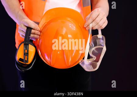Individual in orange vest displaying set of protective headgear including hard hat, noise protectors, and eye shields on black background. Stock Photo