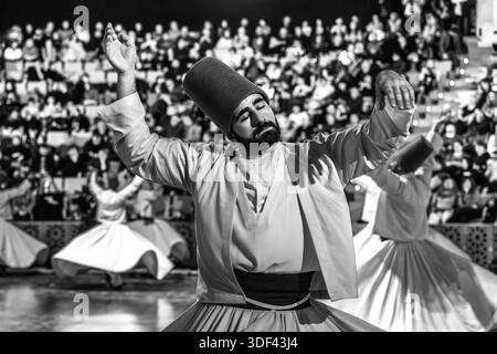The Şeb-i Arus ceremony at the Mevlana Kültür Merkezi in Karatay, Konya, Turkey, a venue that holds up to 3,000 people, shows a bearded dervish with raised arms in the foreground during the ritual Sema dance, his expression calm and inward-turned. The right hand points upwards, receiving God's blessing. The left hand points downwards, passing it on to the earth. The dervishes spin counter-clockwise, symbolically against the ego. The right foot stays fixed while the left foot drives the rotation. Şeb-i Arus is the annual festival marking the death anniversary (17 December) of the mystic poet Ru Stock Photo