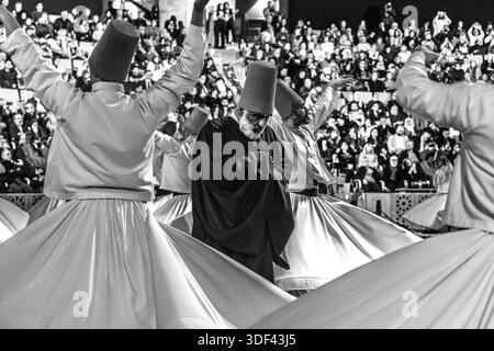 The Şeb-i Arus ceremony at the Mevlana Kültür Merkezi in Karatay, Konya, Turkey, a venue that holds up to 3,000 people, shows the Gözcü (overseer) in a dark robe, his gaze lowered, walking between the widely swinging white skirts of several dervishes during the ritual Sema dance. The right hand points upwards, receiving God's blessing. The left hand points downwards, passing it on to the earth. The dervishes spin counter-clockwise, symbolically against the ego. The right foot stays fixed while the left foot drives the rotation. Among the dancers, the Gözcü usually remains inconspicuous to the  Stock Photo