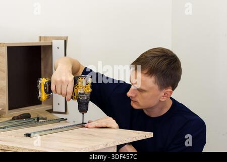 Man screwing drawer rail into cabinet side with power tool. Man carefully screwing metal drawer rail into light wood cabinet side using yellow cordles Stock Photo