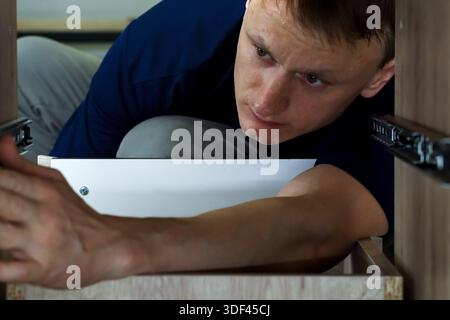Handyman checking alignment of drawer slide in cabinet. Worker carefully aligning and adjusting metal drawer guide during furniture assembly process. Stock Photo