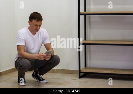 Young man crouching and focused on smartphone screen. Young adult man in casual clothes squatting on floor, intently looking at mobile phone in hands Stock Photo