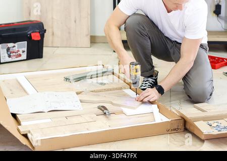 Man crouching and drilling while assembling wooden furniture. Young adult man in white t-shirt squatting on floor using cordless drill to assemble lig Stock Photo