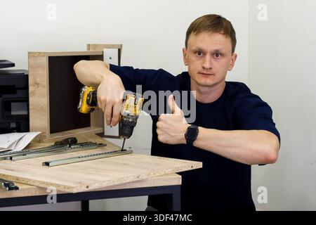 Man showing thumbs up with drill during furniture assembly. Smiling man giving thumbs up approval while holding cordless drill and screwing furniture Stock Photo
