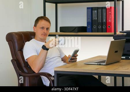 Office worker multitasking with smartphone and coffee cup next to portable computer in comfortable chair setup. (95 characters) Stock Photo