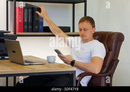 Attentive young professional extends arm to lift red binder from organized shelf in office, glances at smartphone beside laptop on wooden desk. Stock Photo