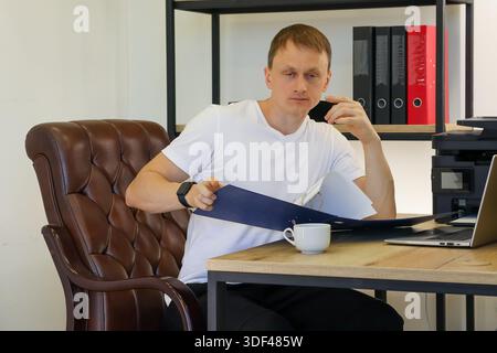Man seated in leather chair holds blue folder, touching chin thoughtfully while reviewing documents in bright office space. Stock Photo