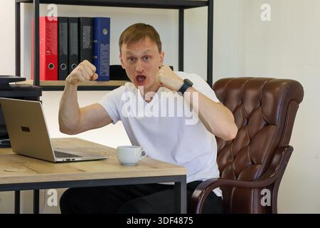Energetic male worker yells triumphantly raising both fists at wooden desk with open laptop in well-lit office with binders Stock Photo