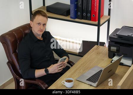 Young professional sits at desk holding phone, facing forward in office with laptop and organizational tools. Stock Photo