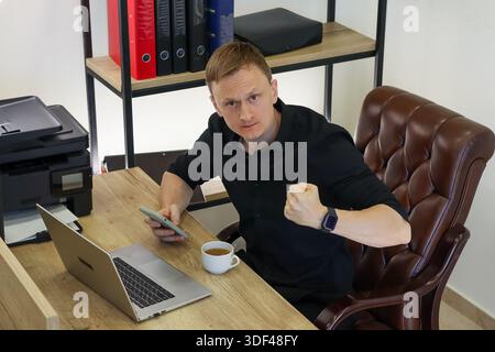 Frustrated worker in suit clenching fist in anger, surrounded by office supplies and tech. Stock Photo