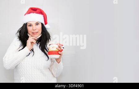 Woman in Santa hat holding festive mug and thinking on white background Stock Photo