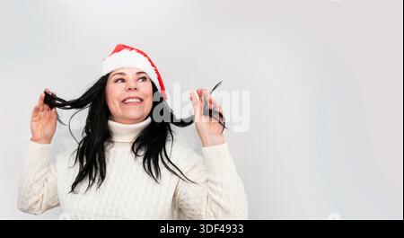 Woman in Santa hat smiling and holding hair wearing white sweater indoors Stock Photo