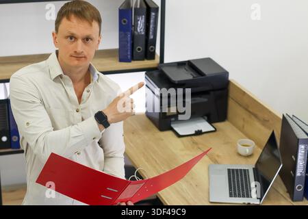 Professional in light shirt extends finger to highlight information in red folder at workstation with printer and computer nearby. Stock Photo