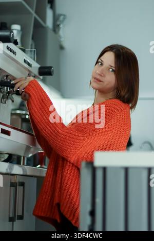 A casual young woman with brown hair wearing an orange knit sweater pulls a lever on a white espresso machine in a bright cafe kitchen setting. Stock Photo