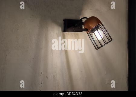 Angled view of industrial wall lamp on textured surface, brown shade over wire cage enclosing glowing bulb. Stock Photo