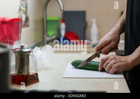 Hands holding a knife to slice a green cucumber on a cutting board, next to a sink with a metal teapot and clear plastic bags on tiled counter. Stock Photo