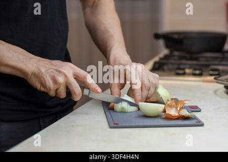 A man in black t-shirt slices white onions using a sharp knife on a gray cutting board placed on a kitchen countertop near the stove. Stock Photo