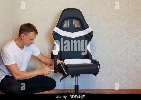 A man in white shirt installs a white cushion headrest onto the arm of a black gaming chair while kneeling in a room. Stock Photo