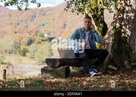 A young man relaxes on a log bench by a large tree, chatting on his phone with an open laptop nearby in a vibrant autumn forest with mountains, river, Stock Photo