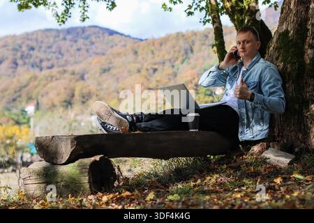 Casual young man with laptop and coffee sits against tree trunk in fall woodland, making thumbs up gesture during call. Stock Photo