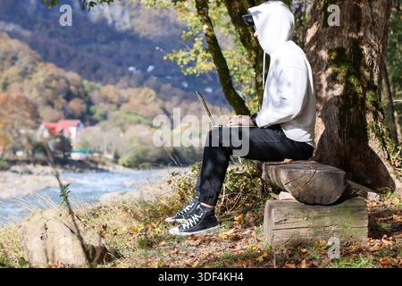Male in hooded sweatshirt operates laptop while resting on stump by tree in fall valley with mountain vista Stock Photo