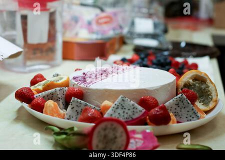 Close-up of fresh strawberries, dragon fruit, passion fruit, and oranges arranged around a white dessert with purple topping on a white plate Stock Photo