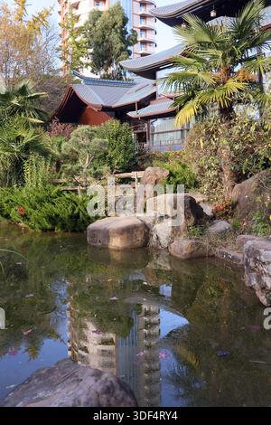 Traditional pagoda structure with blue tiled roofs and modern glass elements beside a rocky pond in a lush garden setting. (118 characters) Stock Photo