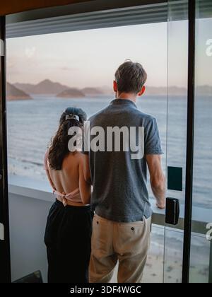 Rear view of a couple standing on a balcony looking out to the ocean at sunset. Stock Photo