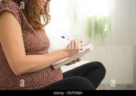 Pregnant woman making to-do lists in a notebook. Calm moment of preparation at home. Stock Photo