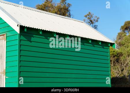 A bright green wooden beach hut featuring a corrugated metal roof and a door, set against a clear blue sky. Stock Photo