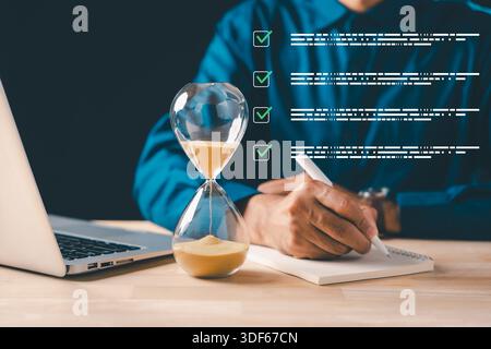 Business professional using pen to write notes in book, hourglass showing passing time, laptop nearby, symbolizing task completion, project planning, Stock Photo