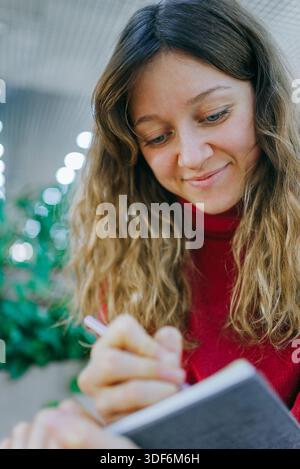 Young caucasian woman in a red turtleneck, curly hair, smiling as she writes in a small notebook with a pen, focused and content while journaling or planning ideas Stock Photo