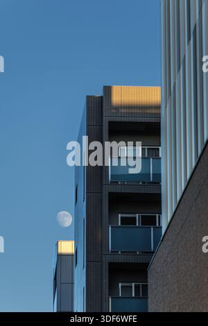 Full moon appears in blue sky over buildings catching last warm light of day as they disappear into shadows of surrounding buildings during sunset. Stock Photo
