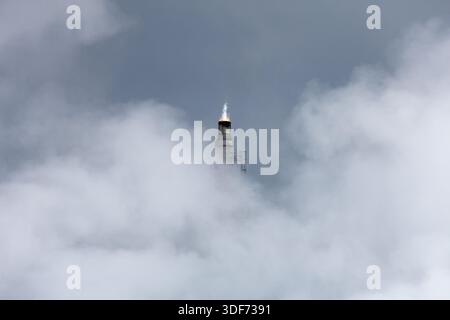 Lone flare stack shrouded in vapor clouds emits flame with sharp white plume stretching upward against background of an errie overcast gray-blue sky. Stock Photo