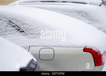 A white car parked on a snowy street, viewed from the rear, completely covered in snow with a red taillight visible. Stock Photo