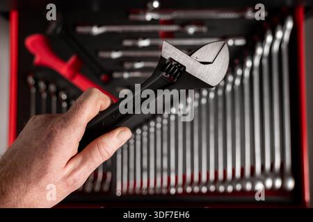 Hand holding adjustable wrench above an organized tool drawer, with neatly arranged metal tools and workshop equipment in the background Stock Photo