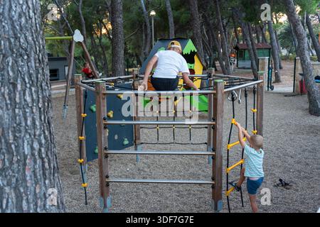 Two brothers playing on climbing frame with rope ladder and bars in resort playground among pine trees in summer evening. Concept of active siblings, Stock Photo