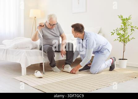 Young nurse or health care worker helping a senior patient with crutches to put on shoes at home. Stock Photo